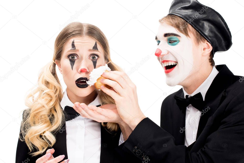 cheerful clown throwing cupcake in face of surprised woman isolated on white 