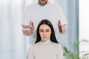cropped shot of bearded healer doing reiki therapy session to young woman with closed eyes clipart