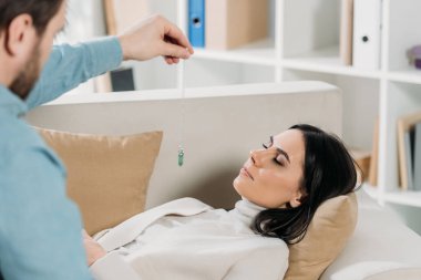 cropped shot of bearded hypnotist hypnotising young woman with closed eyes lying on couch clipart