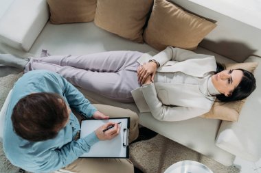 overhead view of psychotherapist writing on clipboard and upset young patient lying on couch clipart