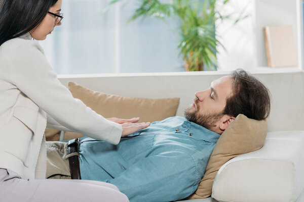 вид на бородатого мужчину с закрытыми глазами лежащего на диване и получающего лечение рейки на груди
