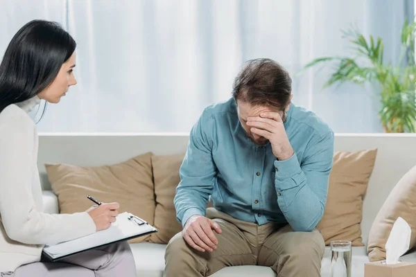 Joven Psicoterapeuta Escribiendo Portapapeles Mirando Hombre Barbudo Molesto Sentado Sofá — Foto de Stock