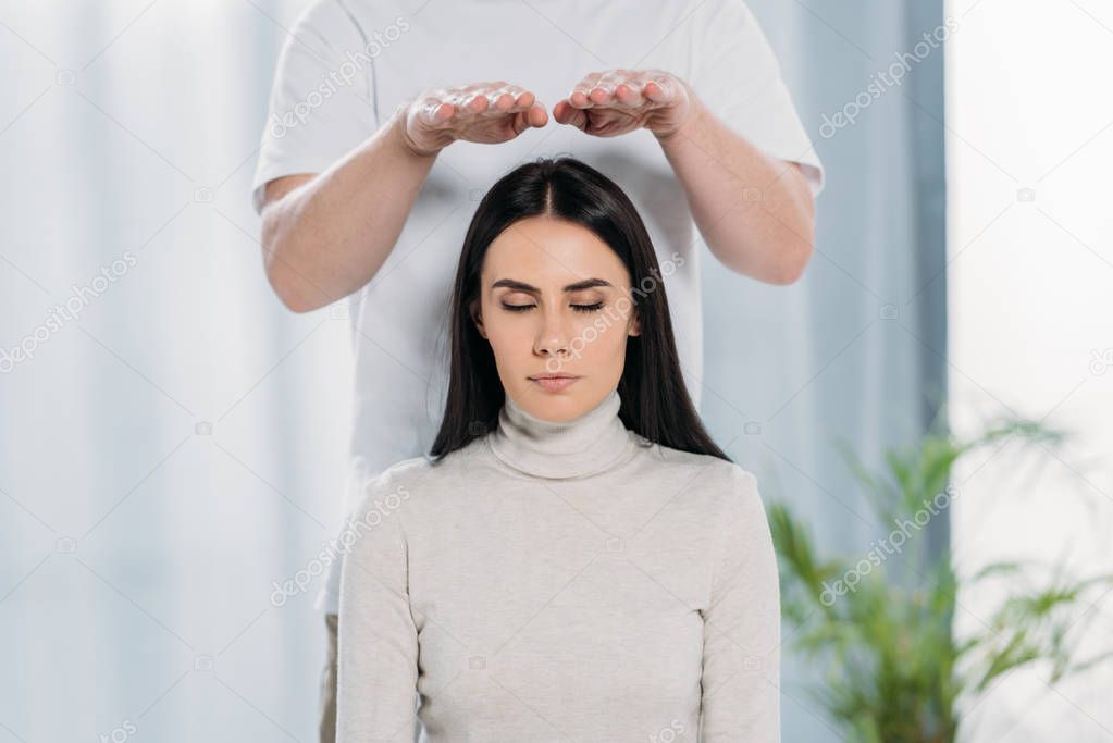 calm young woman with closed eyes receiving reiki healing treatment above head 