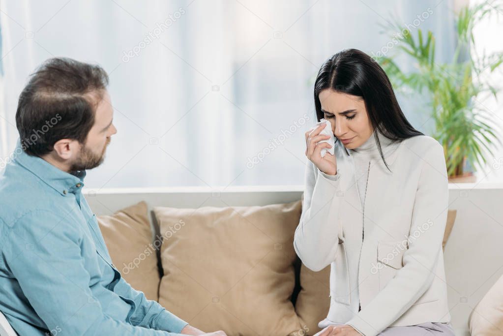 professional psychotherapist looking at young female patient crying on couch
