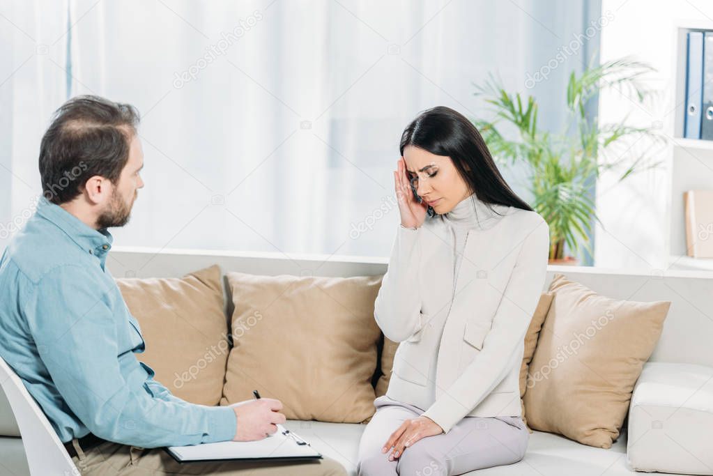 unhappy young woman with depression sitting on couch and psychiatrist writing on clipboard