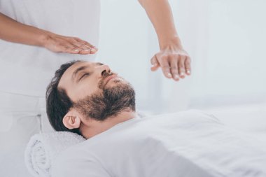 calm bearded man lying on massage table and hands of healer doing reiki treatment session clipart