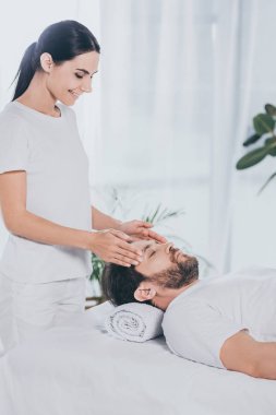 smiling young woman doing reiki treatment session to calm bearded man with closed eyes clipart