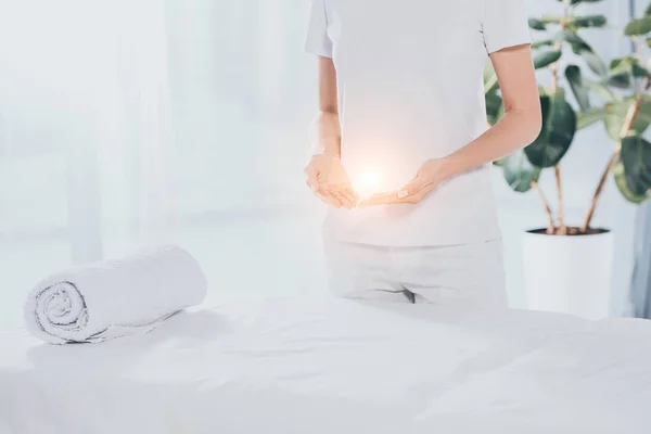 mid section of reiki healer with light energy in hands standing near white massage table