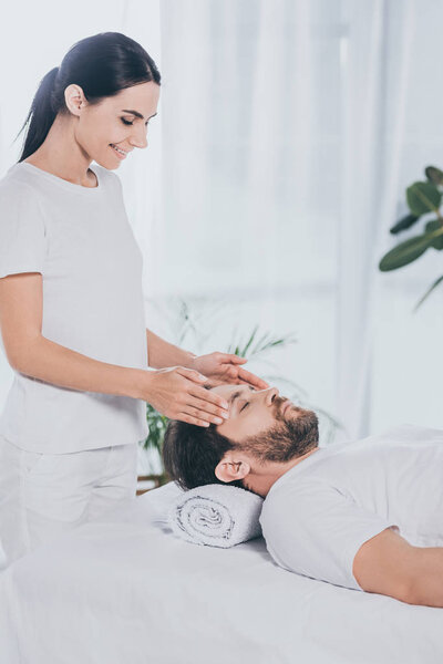 smiling young woman doing reiki treatment session to calm bearded man with closed eyes