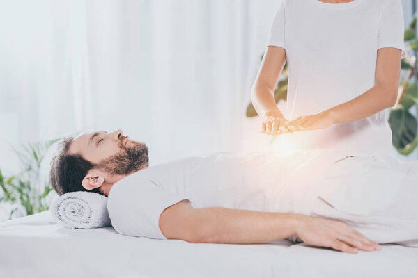 calm bearded man with closed eyes lying on massage table and receiving reiki treatment