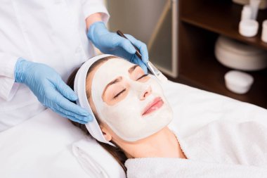 beautician standing and holding cosmetic brush near woman with cosmetic mask on face at beauty salon clipart