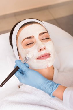 beautician applying cosmetic mask to woman on face with cosmetic brush at beauty salon clipart