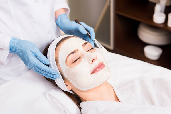 beautician standing and holding cosmetic brush near woman with cosmetic mask on face at beauty salon