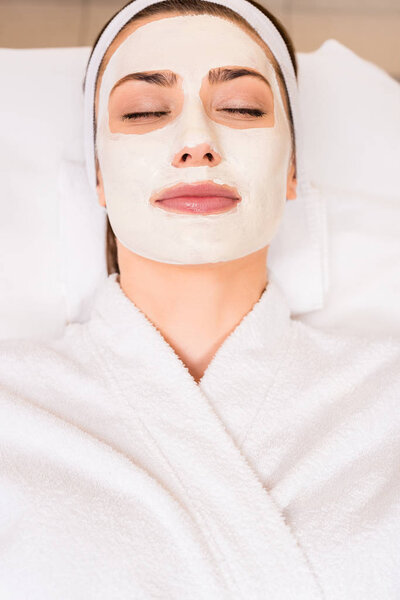 top view of woman lying in white bathrobe with applied face mask at beauty salon