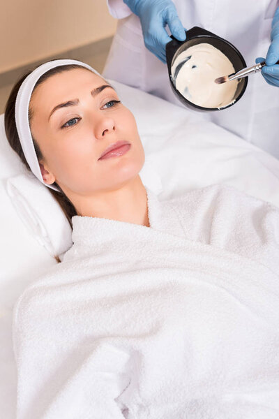 beautician mixing face mask while woman lying on bathrobe and hairband at beauty salon