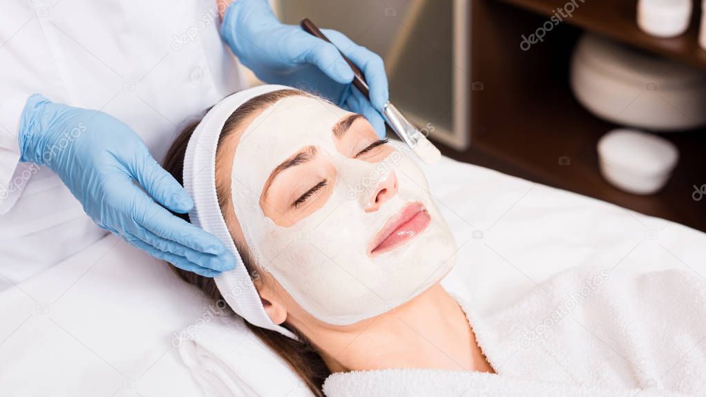 beautician standing near woman with cosmetic mask on face and holding cosmetic brush at beauty salon