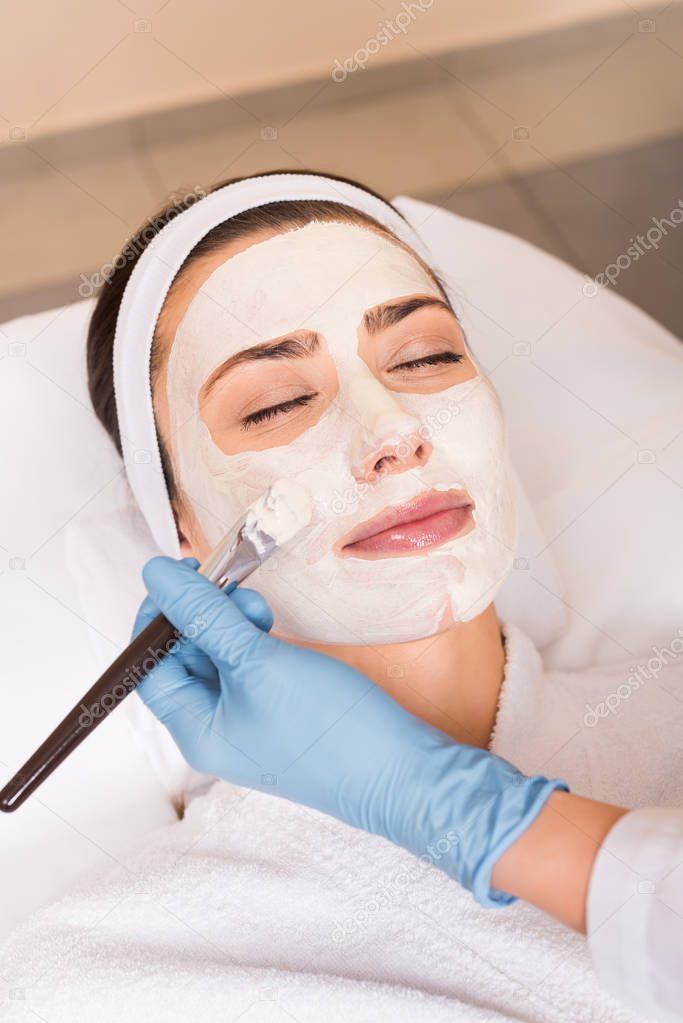 beautician applying cosmetic mask to woman on face with cosmetic brush at beauty salon