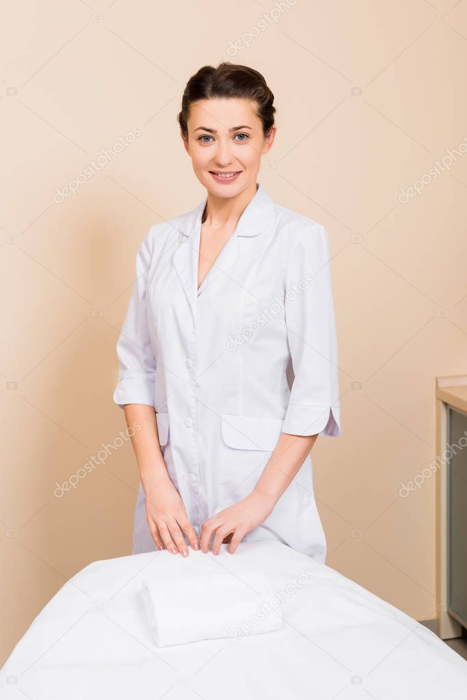 beautician standing behind massage table and smiling at beauty salon