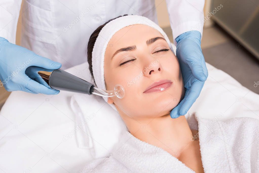 beautician doing darsonvalization with equipment to woman on cheek at beauty salon