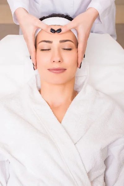 バスローブ 美容院でヘアバンドに横たわる女に手動フェイス マッサージをやっている美容師 — ストック写真