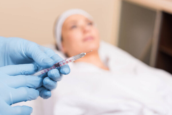 selective focus of syringe in beautician hands in front of woman face at beauty salon