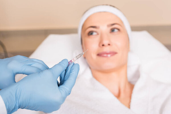 selective focus of syringe holding by cosmetologist over woman face at beauty salon