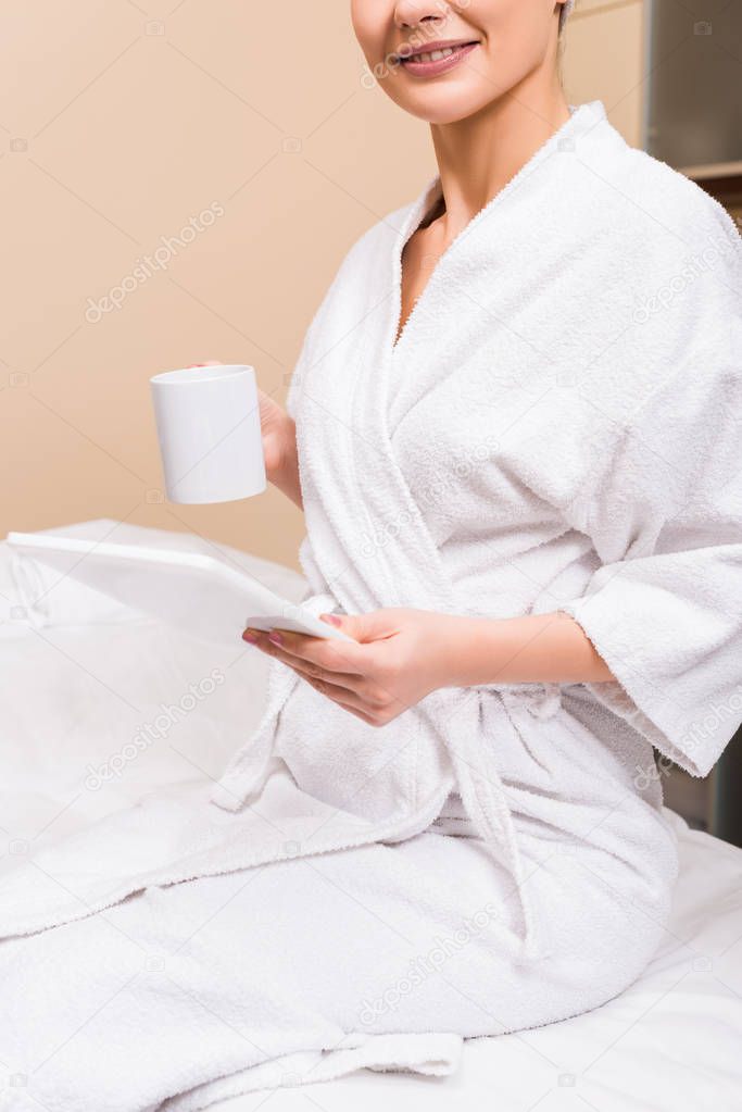 cropped view of woman sitting, holding cup and digital tablet at beauty salon