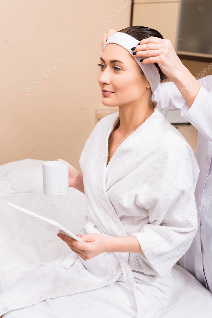 woman sitting, holding cup and using digital tablet while beautician correcting her hairband at beauty salon