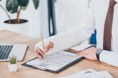 cropped view of worker with broken arm in gypsum sitting at table and filling out document in office, compensation concept clipart