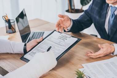 cropped view of worker with broken arm signing form for compensation claim  opposite to businessman in blue jacket in office, compensation concept clipart