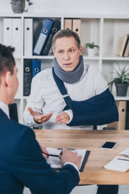 middle aged worker in neck brace with broken arm sitting at table and talking to opposite businessman in office, compensation concept clipart