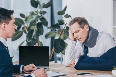 upset worker in neck brace with broken arm sitting at table opposite businessman in blue jacket in office, compensation concept clipart