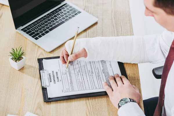 cropped view of worker with broken arm in gypsum sitting at table and filling compensation form in office, compensation concept