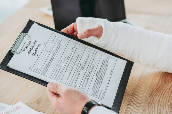 cropped view of worker with broken arm in gypsum sitting at table and holding compensation form in office, compensation concept