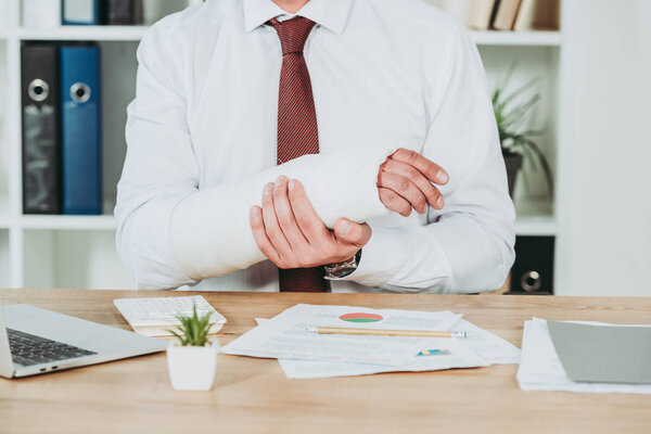cropped view of worker holding broken arm wile sitting at table with documents in office, compensation concept