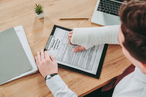 cropped view of worker holding broken arm compensation claim and sitting at table in office, compensation concept