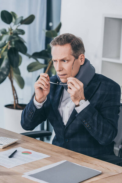 businessman in neck brace sitting at table wiht documents and puting on glasses in office, compensation concept