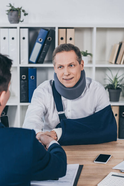 businessman in blue jacket shaking hands with worker in neck brace and arm bandage over table in office, compensation concept