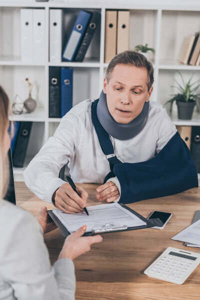 worker in neck brace and arm bandage signing while woman holding paper tablet over table in office, compensation concept