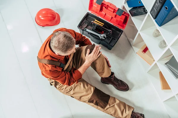 top view of repairman sitting on floor and holding injured knee surrounding by equipment in office