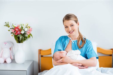 yatakta oturmuş ve yeni doğan bebek emzirme sırasında kamera gülümseyen mutlu genç anne 