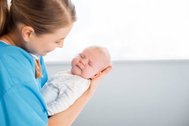 side view of happy young mother carrying adorable sleeping baby in hospital room clipart