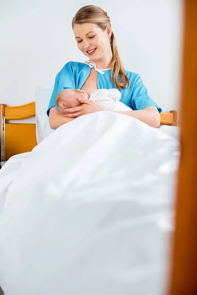 selective focus of beautiful happy young mother breastfeeding newborn baby on bed in hospital room
