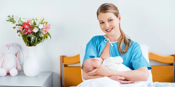 happy young mother sitting in bed and smiling at camera while breastfeeding newborn baby in hospital room