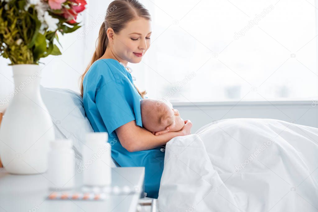 side view of happy young mother sitting on bed and breastfeeding newborn baby in hospital room