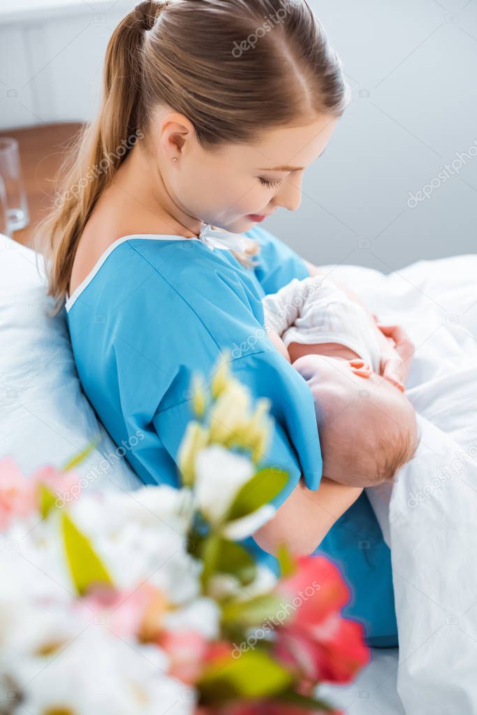 side view of smiling young mother breastfeeding newborn baby on bed in hospital room 