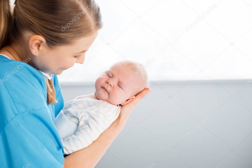 side view of happy young mother carrying adorable sleeping baby in hospital room