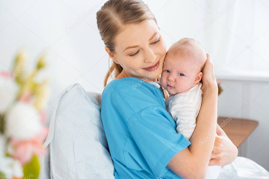 happy young mother sitting on hospital bed and hugging adorable baby 
