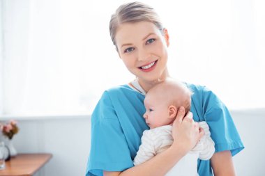happy young mother holding newborn baby and smiling at camera in hospital room clipart