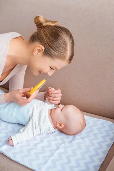 Çıngırak Tutarak Koltukta Yalan Wth Bebek Oynarken Mutlu Genç Anne — Stok fotoğraf